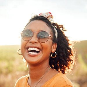 A young woman smiling confidently outdoors while wearing sunglasses.